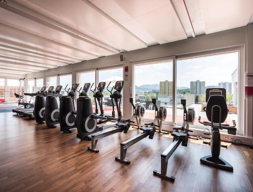 Fitness center with cardio machines lined up against large windows overlooking the city.